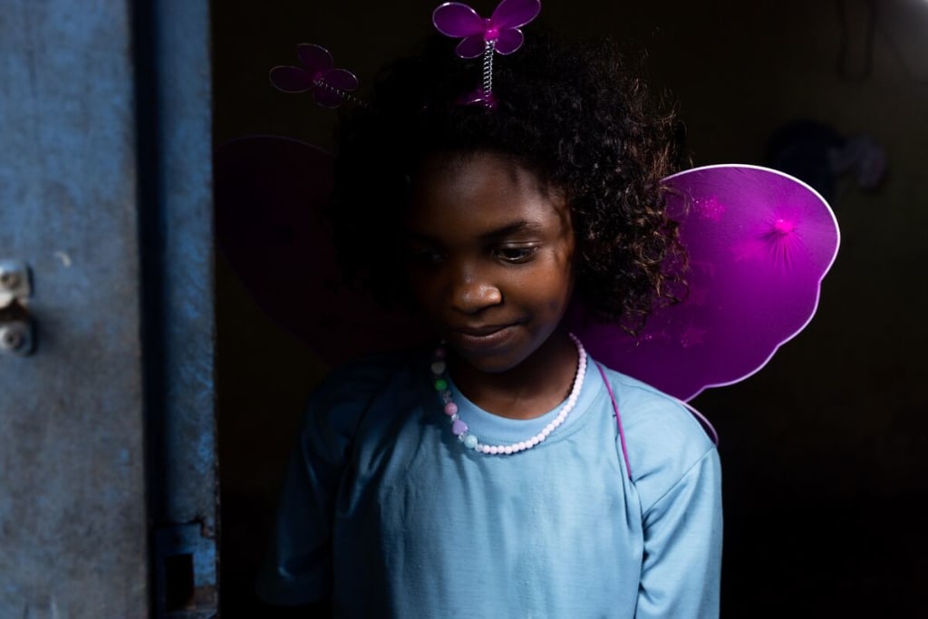 A girl in a blue shirt looks down and wears pink wings on her back